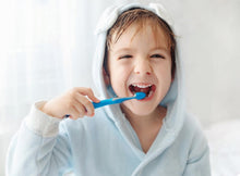 Charger l&#39;image dans la galerie, Child brushing teeth with blue toothbrush, smiling in cozy bathrobe, promoting healthy dental hygiene.